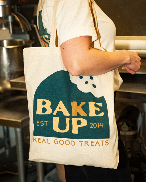 Image of a subject carrying a tote back on their shoulder. The bag has the company logo on it. The logo is a green dome shape with a bite taken out of it in the upper right hand corner. The Text embedded within the logo reads "Bake Up" and "Est. 2014" in a mix of cream and orange colors. The text below the green dome shape reads "Real good treats" in all-caps and orange. Background is of an industrial kitchen.