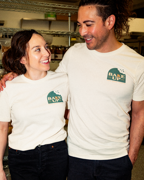 Image of a smiling couple, looking at each other in matching T-shirts. The male in the photo is on the right hand side with has left arm around the female's shoulder . The female subject is on the left hand side with her right arm behind his back. The T-shirt is white with the company logo in the upper right hand corner. Logo is green with the text "Bake Up", "Est. 2014", and "Real good treats" in orange and cream colors. Background is of industrial kitchen.