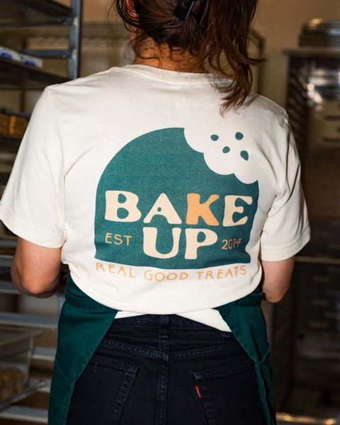 Female, showing back of T-shirt where the logo is large and centered. The logo is a green half circle with a bite taken out of the top right side. Nestled inside the shape is the Company name "Bake Up" with the words "Est. 2014". At the bottom is the tagline "Real Good Treats" in all caps and orange text. Background is industrial kitchen.