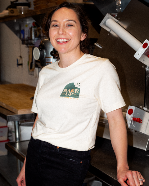 Female smiling and wearing the white Bake Up T-shirt while standing in an industrial kitchen.