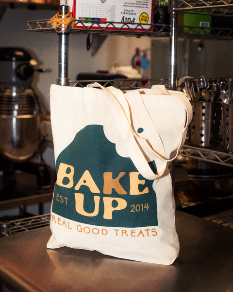 Image of the tote bag on it's own, sitting on an industrial kitchen counter. The bag is white with the company's logo which is a green dome shape with a bite taken out of it in the upper right hand corner. The Text embedded within the logo reads "Bake Up" and "Est. 2014" in a mix of cream and orange colors. The text below the green dome shape reads "Real good treats" in all-caps and orange.