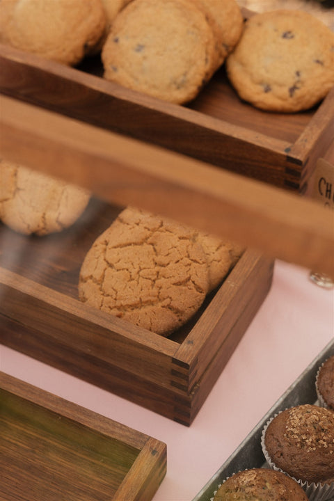 Camera peaks over the display case to see more pastry trays along the table. Various pastries pictured which include chocolate chip cookies, peanut butter cookies, and zucchini muffins. 